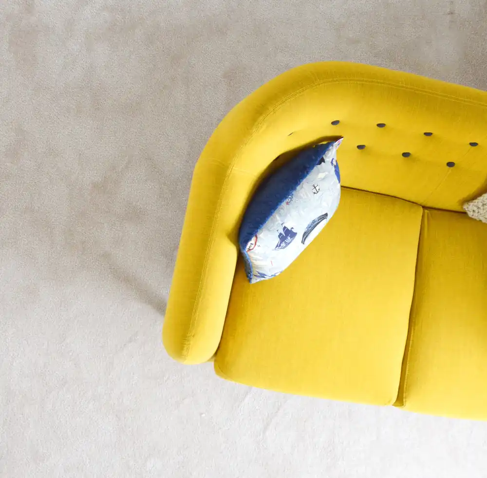 Vue de dessus d'un canapé jaune vif posé sur un tapis crème, avec un coussin à motif bleu et blanc.