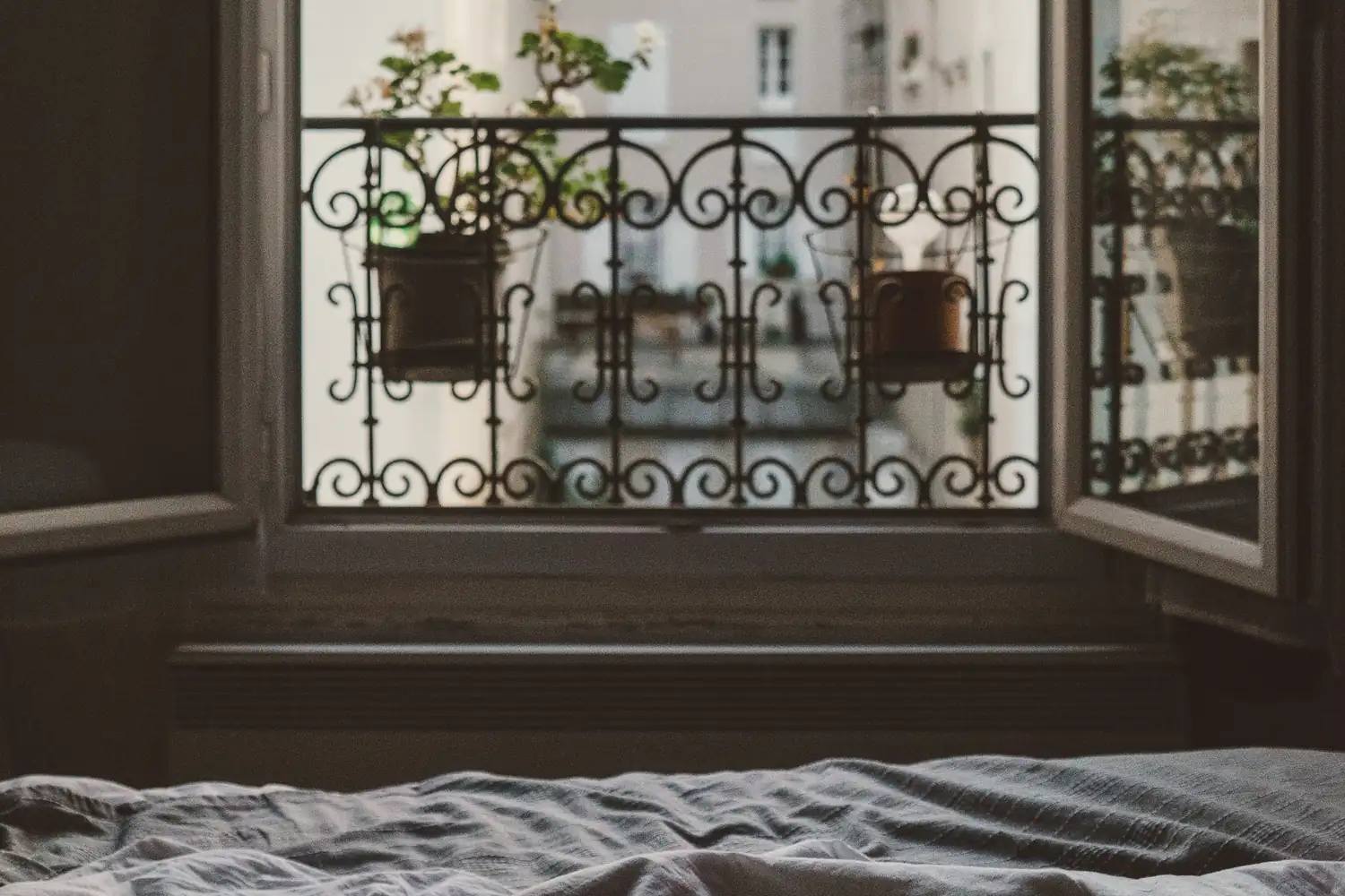 Une chambre dont la fenêtre à deux battants est grande ouverte sur un balcon en fer forgé garni de plantes ; au premier plan, un lit défait aux draps de lin gris.