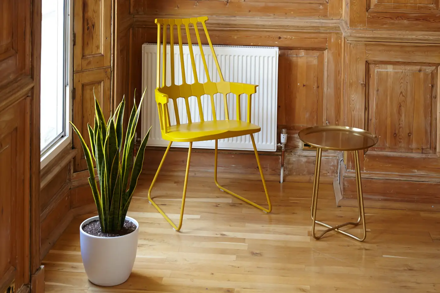 Un angle de bureau lambrissé de bois clair : une chaise jaune vif devant le radiateur, une plante verte en pot blanc et un guéridon rond en laiton.