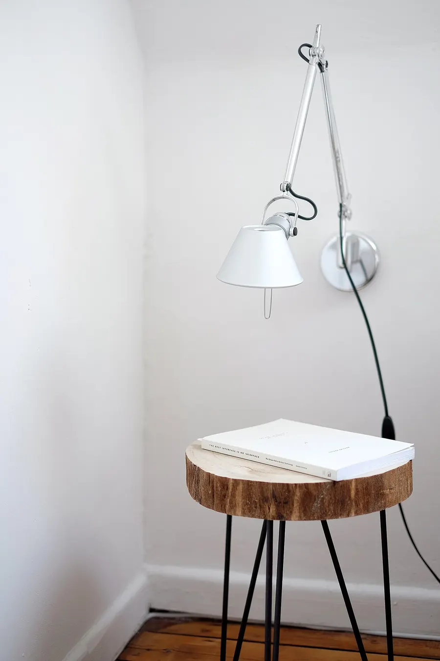 Une lampe d'architecte blanche fixée au mur, au-dessus d'une petite table faite d'un rondin de bois posé sur trois pieds fins.