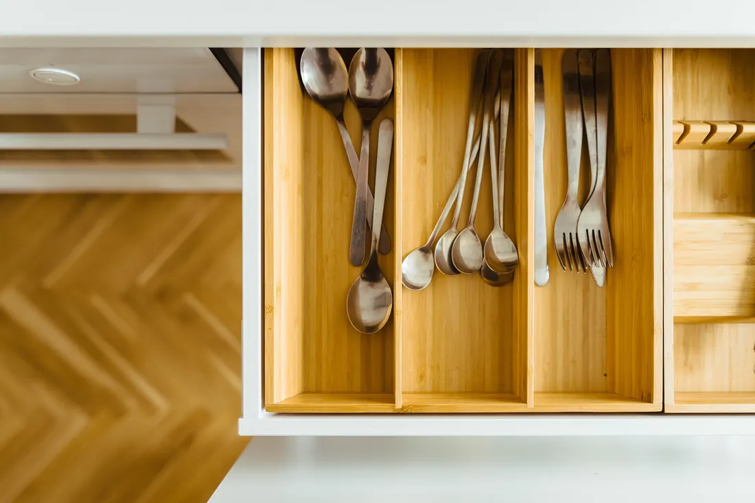 Un tiroir de cuisine ouvert, compartimenté en bois clair, où les couverts sont rangés par famille au-dessus d'un parquet à chevrons.