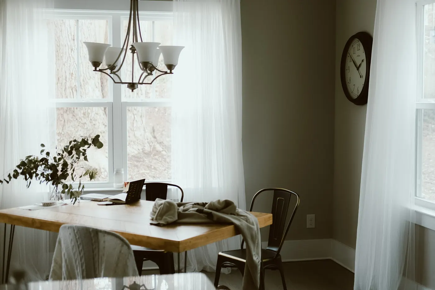 Une salle à manger claire : table en bois clair devant une grande fenêtre voilée de rideaux blancs, chaises en métal noir, lustre à cinq branches et branchages d'eucalyptus dans un vase.