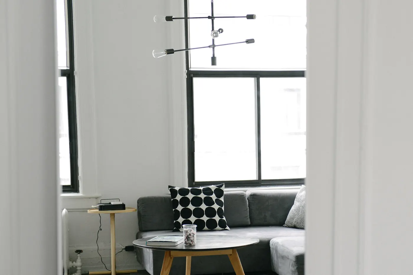 Un coin salon aperçu par l'embrasure d'une porte : canapé d'angle gris, table basse ronde en bois et suspension à trois branches devant une haute fenêtre.