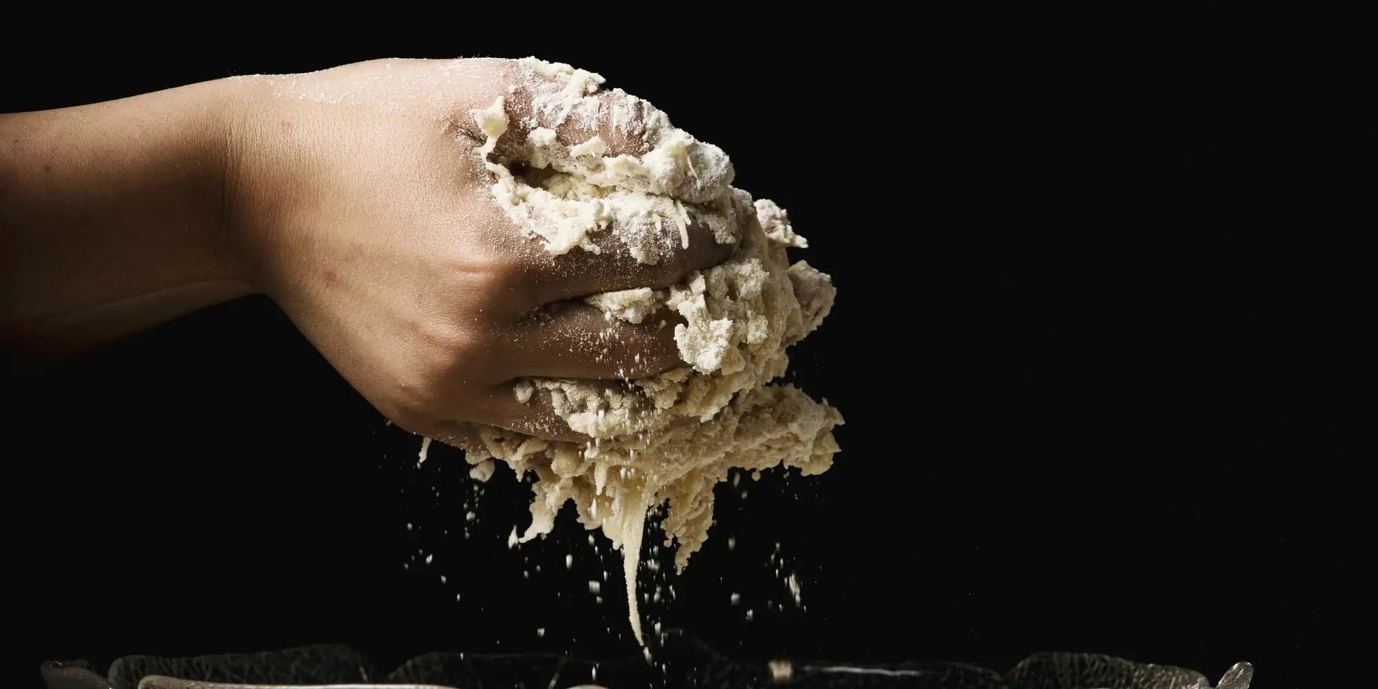 Une main serre une poignée de pâte farinée au-dessus d'un saladier, sur fond noir.